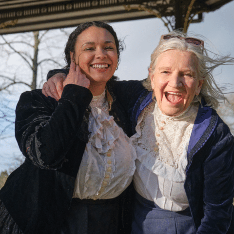 Adaya Henry and Janice Connolly in victoria period costumes laughing and hugging at the bandstand in Cannon Hill Park