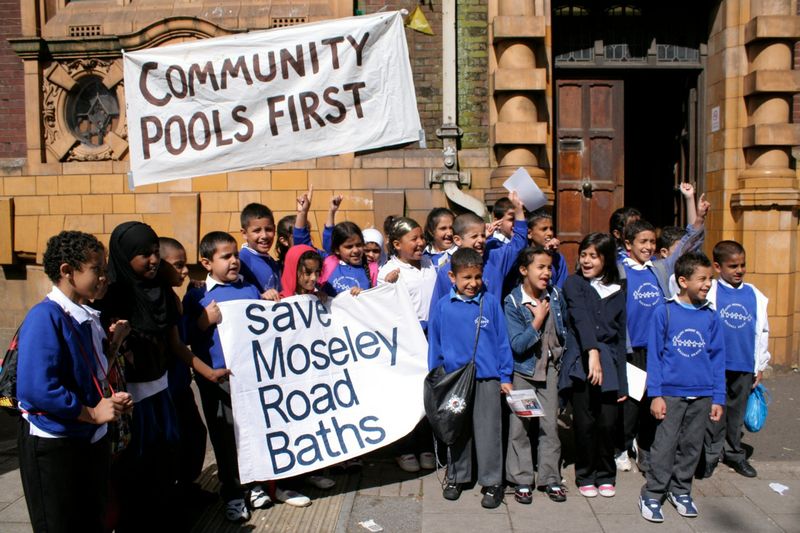 A group of school children outside moseley road baths with banners saying community pools first and save moseley road baths