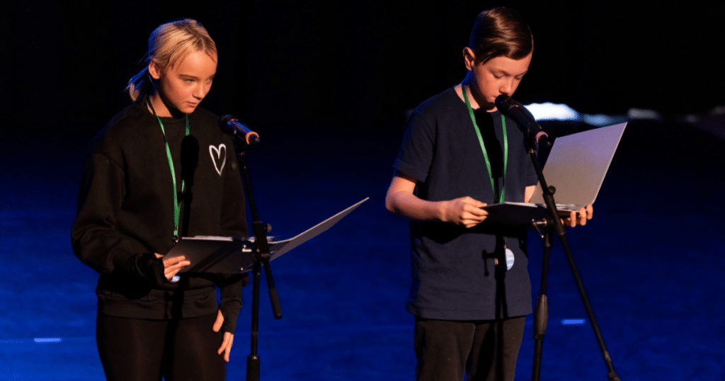 2 young people reading poetry on a stage into microphones